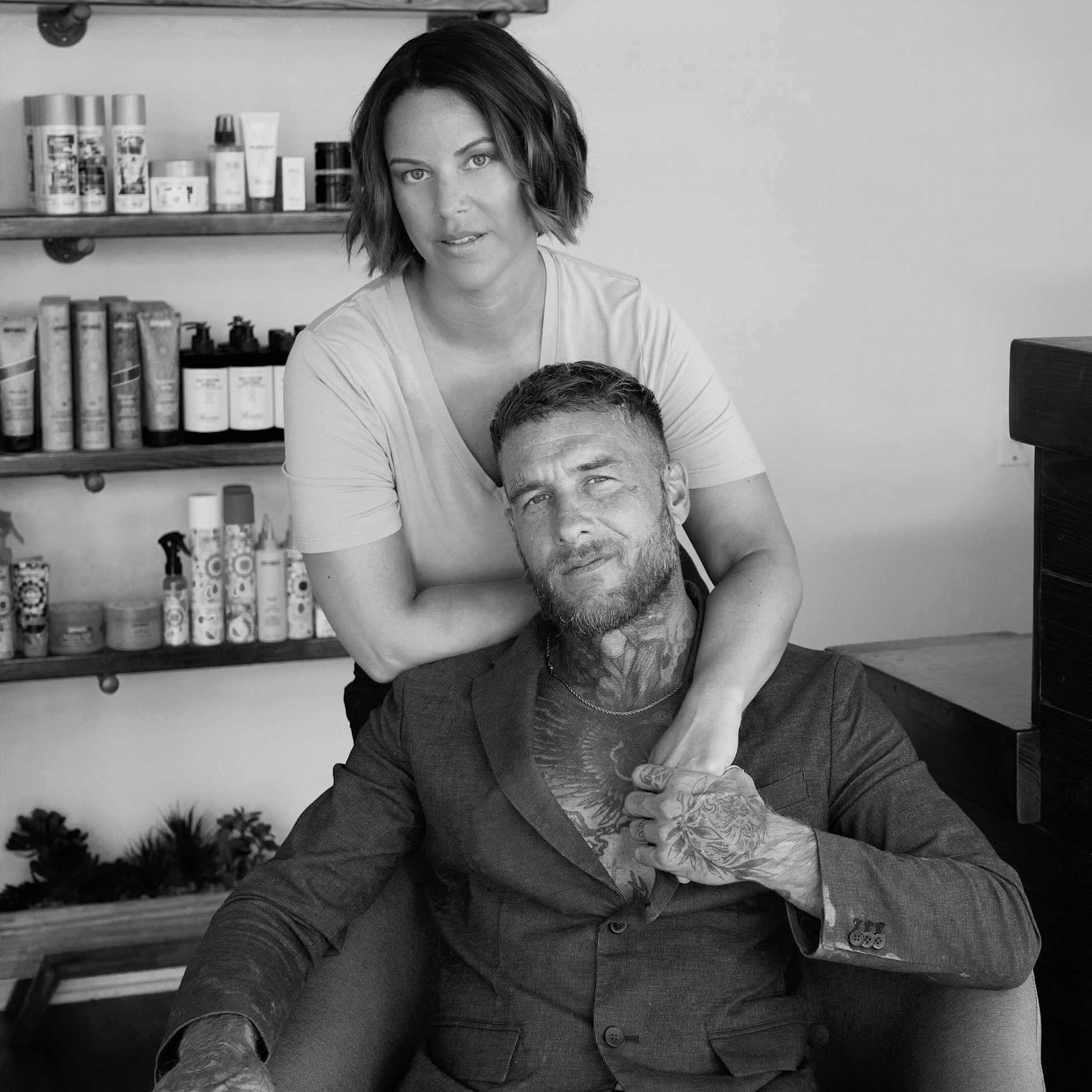 Woman standing behind a seated man in a suit with shelves of hair products in the background.