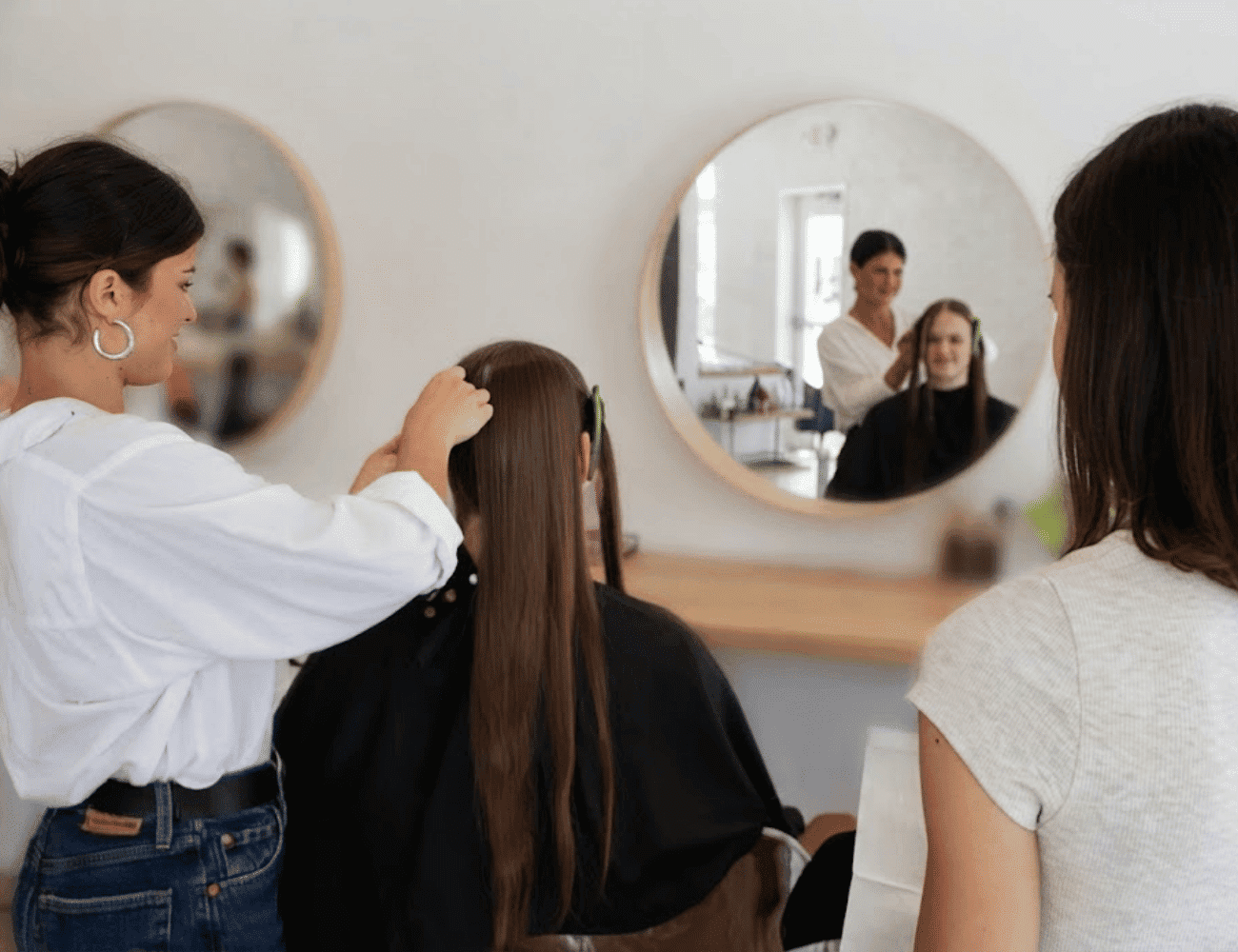 Hairdresser styling a woman's long hair in a salon, with reflections in round mirrors.