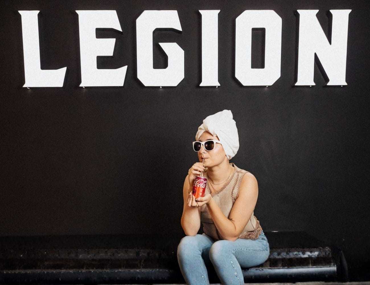 Woman in sunglasses sips a drink beneath a large "LEGION" sign, seated on a bench.