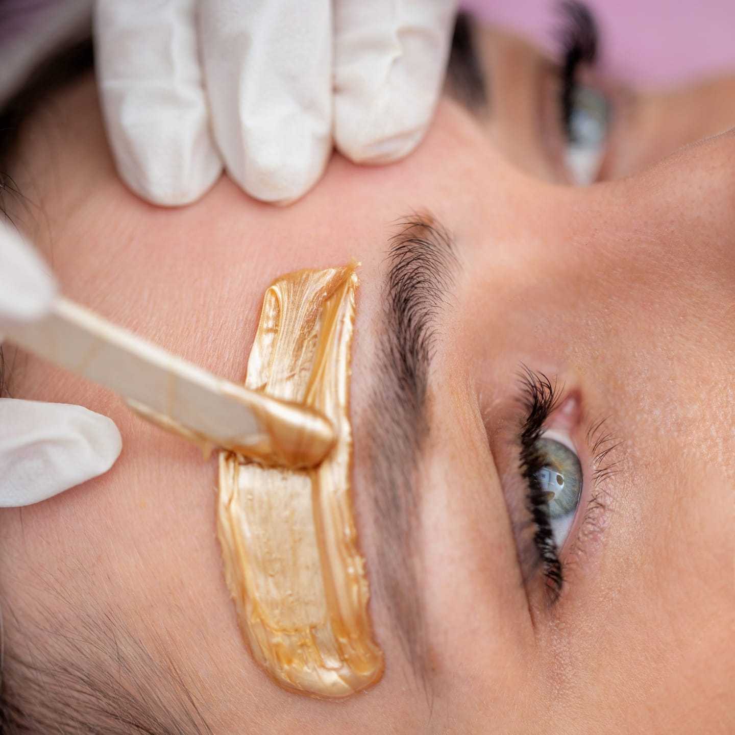 Person receiving an eyebrow waxing treatment with gold-colored wax applied above the eyebrow.