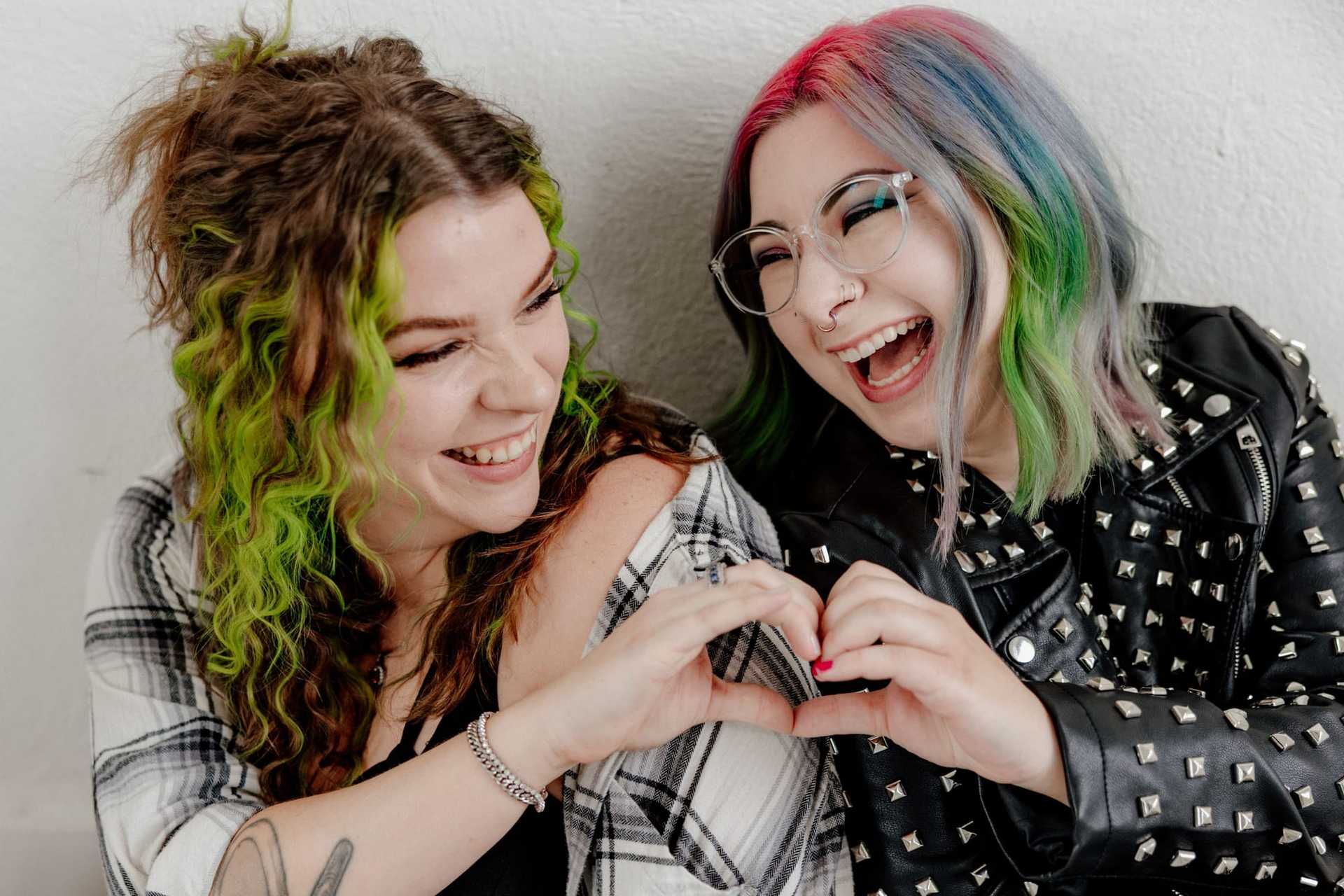 Two friends with colorful hair making a heart shape with their hands, smiling joyfully.