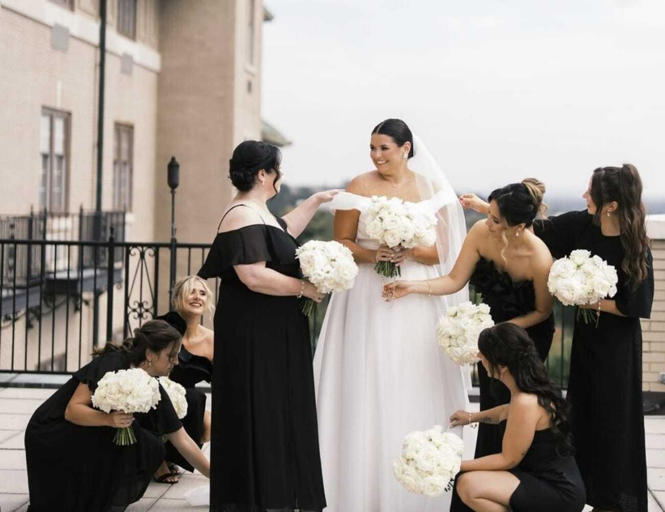 Bride with bridesmaids in black dresses, holding white bouquets on a terrace.