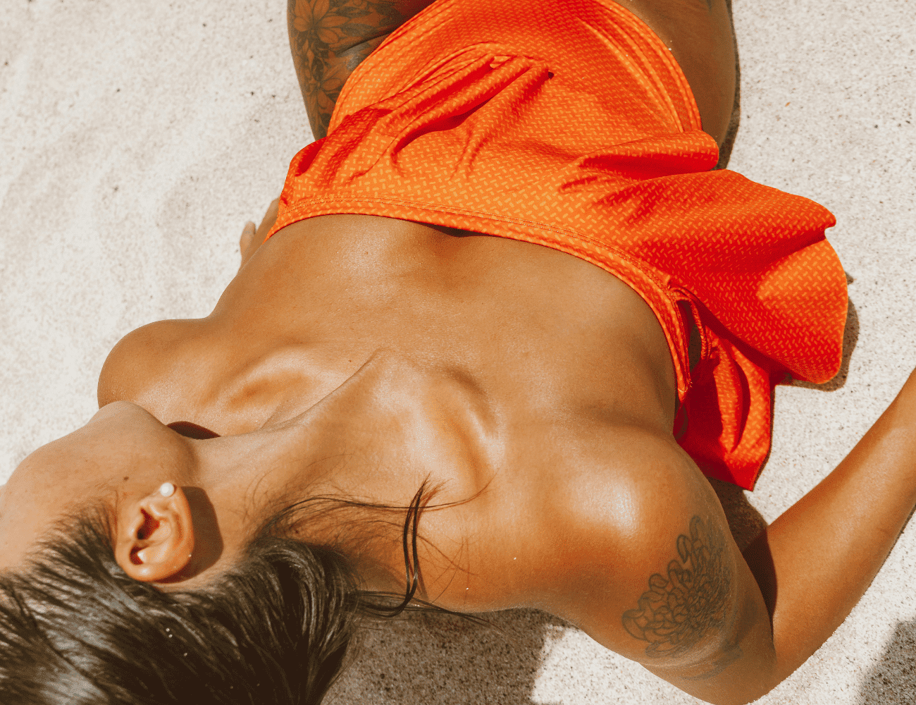 Person in bright orange swimsuit relaxing on sandy beach, showing shoulder tattoos.